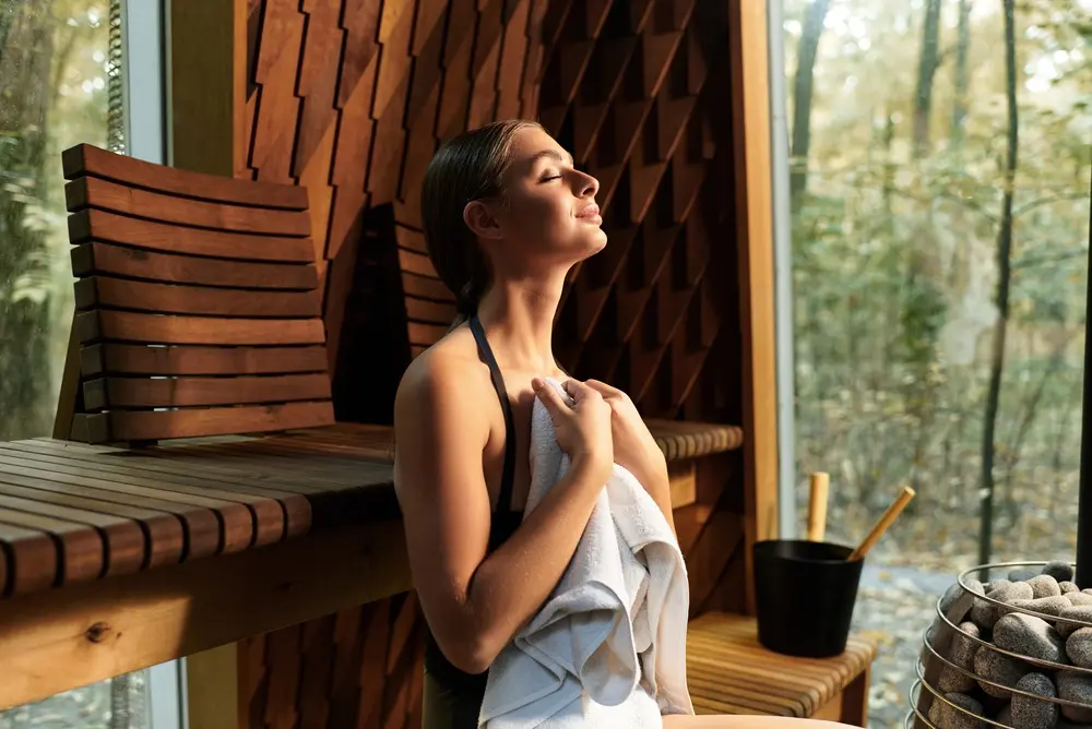 Femme détendue, les yeux fermés et une serviette contre elle, profitant de la chaleur d'un sauna en bois lors d'un week-end bien-être