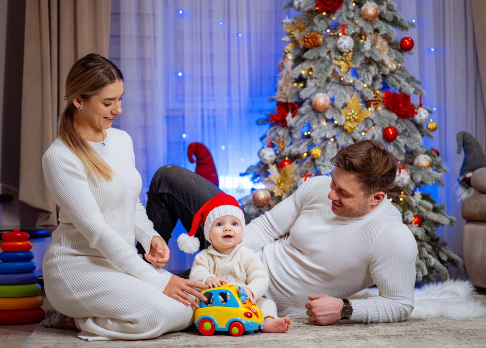 Parents jouant avec leur bébé devant un sapin de Noël, réunis pour découvrir leurs cadeaux