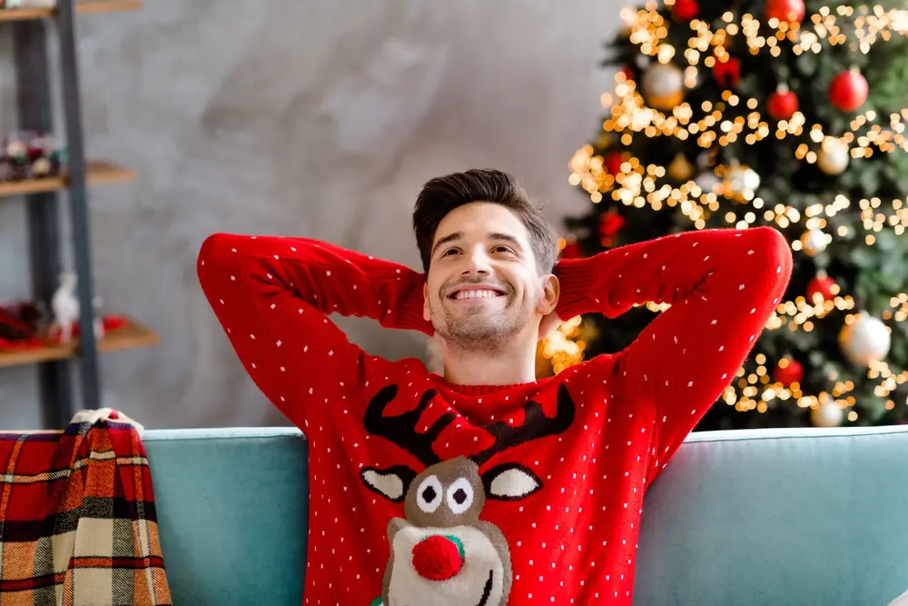 Homme souriant en pull de Noël, détendu sur son canapé avec un sapin illuminé