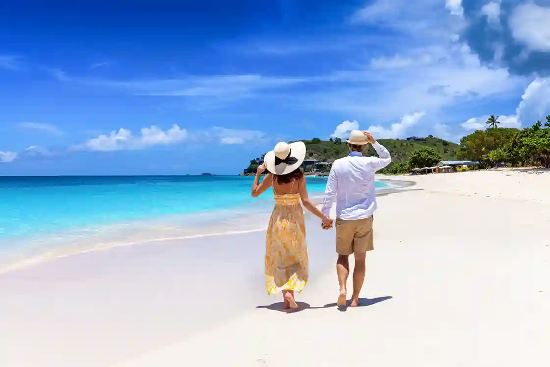 Un couple en lune de miel, coiffé de chapeaux de soleil, se promène sur une plage tropicale bordée par une mer turquoise dans les Caraïbes, sur l'île d'Antigua.